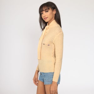 May include: A pale yellow ribbed knit sweater with a tie at the neck. The sweater has a small embroidered design on the front.
