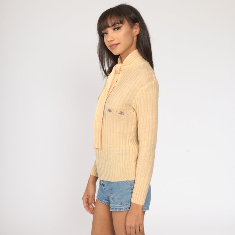 May include: A pale yellow ribbed knit sweater with a tie at the neck. The sweater has a small embroidered design on the front.