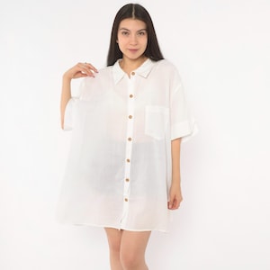 May include: A crisp white button-down shirt featuring a classic collar, short sleeves, and a single chest pocket. The garment is constructed with a lightweight, semi-sheer fabric and includes a full-length front button placket with brown buttons.