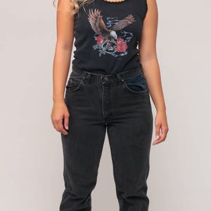 May include: Black tank top with an eagle graphic, paired with dark wash jeans. The tank top features a detailed illustration of an eagle with roses and a skull. The jeans have a classic fit.