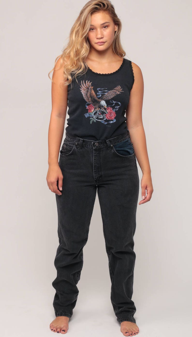 May include: Black tank top with an eagle graphic, paired with dark wash jeans. The tank top features a detailed illustration of an eagle with roses and a skull. The jeans have a classic fit.