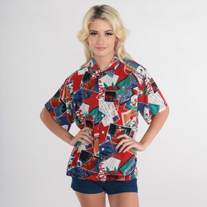 May include: A red, blue, and white short-sleeved button-down shirt with a Hawaiian print featuring the word "Aloha" and images of palm trees, surfers, and postcards. The shirt has a collar and a button closure.