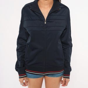 May include: A navy blue zip-up track jacket featuring a high collar, long sleeves, and horizontal stitched detailing across the chest. The cuffs and hem are finished with red and white striped trim. The jacket has a central silver metal zipper.