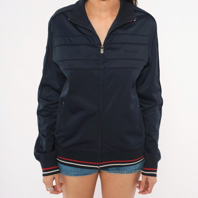 May include: A navy blue zip-up track jacket featuring a high collar, long sleeves, and horizontal stitched detailing across the chest. The cuffs and hem are finished with red and white striped trim. The jacket has a central silver metal zipper.
