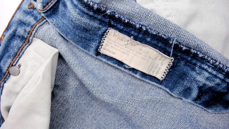 May include: Close-up of blue denim jeans, showing the inside pocket and the Wrangler label. The jeans have a classic, worn look with visible stitching and a metal button. The label provides size and care instructions.