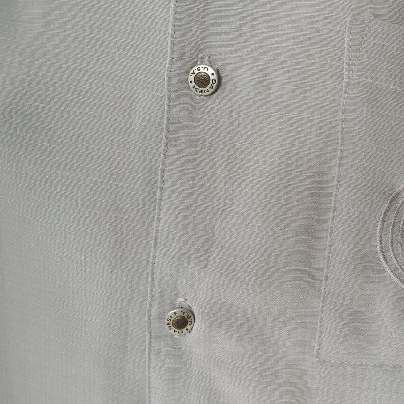 May include: Close-up of a gray button-down shirt with two silver buttons. The buttons have the text "Daniels U.S.A." engraved on them.