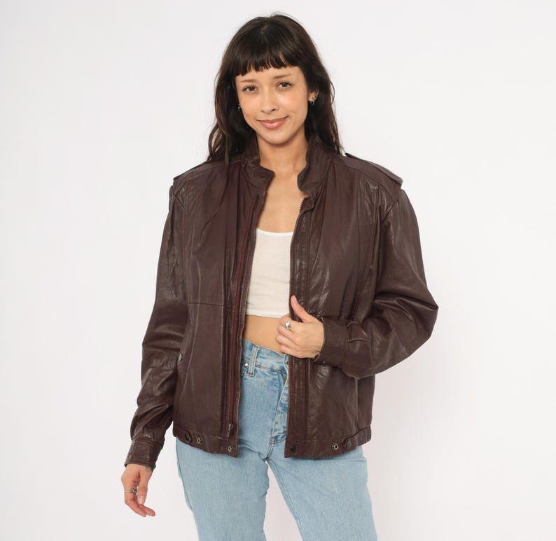 May include: A dark brown leather jacket with a zip-up front, side pockets, and a stand-up collar. The jacket has a slightly worn appearance and is paired with light blue jeans. The model is wearing a white top underneath.