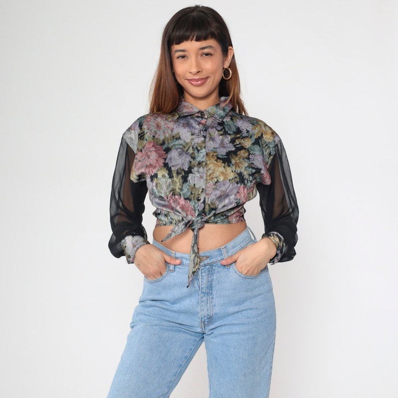 May include: A floral print blouse with sheer black sleeves tied at the waist. The blouse has a button-down collar and a mix of pink, purple, and yellow flowers on a black background. The model is wearing light blue jeans.