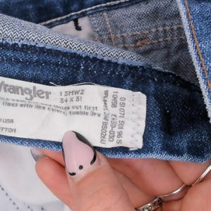 Pode incluir: Close-up de cal&ccedil;as de ganga azuis com uma etiqueta Wrangler. A etiqueta indica um tamanho 34 x 31 e material 100% algod&atilde;o. As cal&ccedil;as de ganga s&atilde;o feitas nos EUA.
