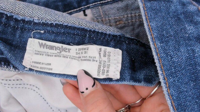 Pode incluir: Close-up de cal&ccedil;as de ganga azuis com uma etiqueta Wrangler. A etiqueta indica um tamanho 34 x 31 e material 100% algod&atilde;o. As cal&ccedil;as de ganga s&atilde;o feitas nos EUA.