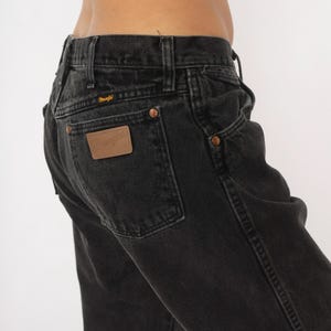 May include: Close-up of black denim jeans with a classic five-pocket design. Features copper rivets and a brown leather patch on the back pocket with the Wrangler logo. The jeans have a faded, worn look.