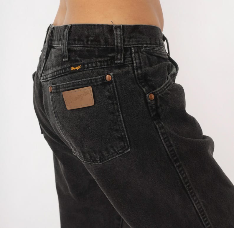 May include: Close-up of black denim jeans with a classic five-pocket design. Features copper rivets and a brown leather patch on the back pocket with the Wrangler logo. The jeans have a faded, worn look.