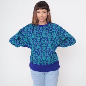 Op de afbeelding: Een blauw en teal patroon trui met een ronde hals en lange mouwen. De trui heeft een herhalend bloemenontwerp en een effen blauwe geribbelde zoom en halslijn. Het model draagt een lichtblauwe jeans.