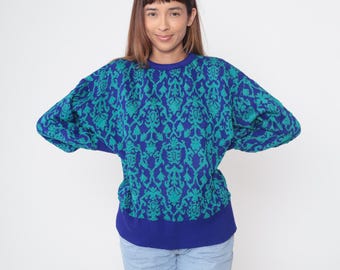 May include: A blue and teal patterned sweater with a crew neck and long sleeves. The sweater features a repeating floral design and a solid blue ribbed hem and neckline. The model is wearing light blue jeans.