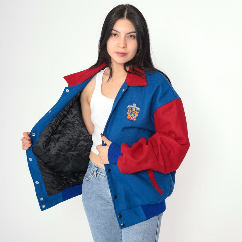 May include: A blue and red varsity jacket with a large embroidered crest on the left side. The jacket has a snap closure and a ribbed collar, cuffs, and waistband.