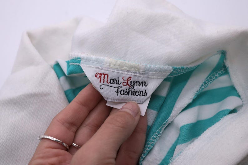 May include: Close-up of a white and turquoise striped shirt with a white tag that reads "Mari Lynn Fashions".