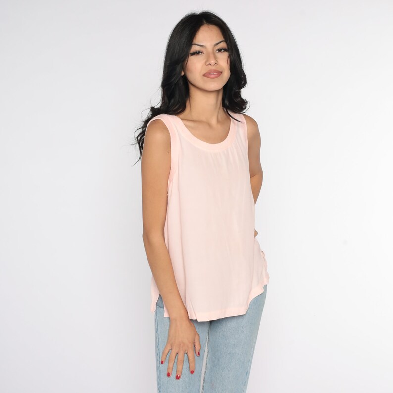 May include: A sleeveless, light pink tank top with a rounded neckline. The top is made of a soft, flowing fabric. The model is wearing light blue jeans. The background is plain white.