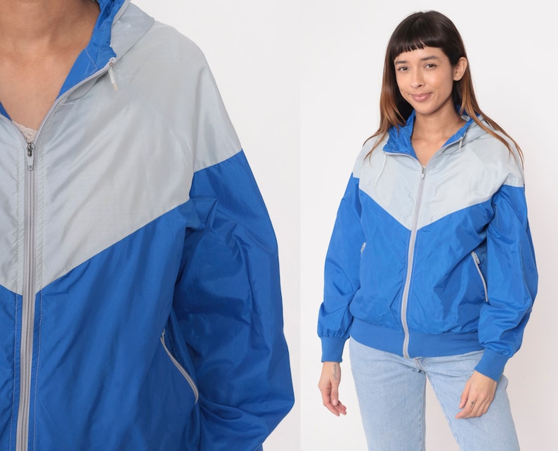 May include: A blue and silver windbreaker jacket with a zipper closure and a hood. The jacket has a color block design with a silver top and a blue bottom.