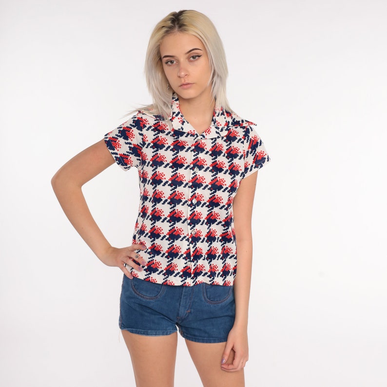 May include: A white, red, and blue patterned short-sleeved button-up shirt with a collared neckline. The shirt is worn with denim shorts.