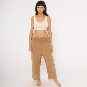 May include: A person wearing a white tank top and tan work pants. The pants have large front pockets and knee patches. The person is standing in front of a white background.