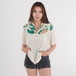 May include: Cream-colored short-sleeve shirt with a V-neck collar. The collar and upper chest feature a green and tan leaf pattern. The shirt is paired with black denim shorts. The model is standing in front of a white background.