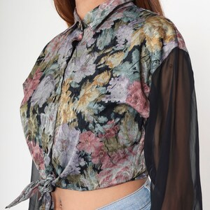 May include: A floral print button-down shirt with black sheer sleeves. The shirt has a tie-front design and a collar. The floral pattern includes shades of pink, purple, green, and gold on a black background. The sleeves are long and made of a sheer black fabric.