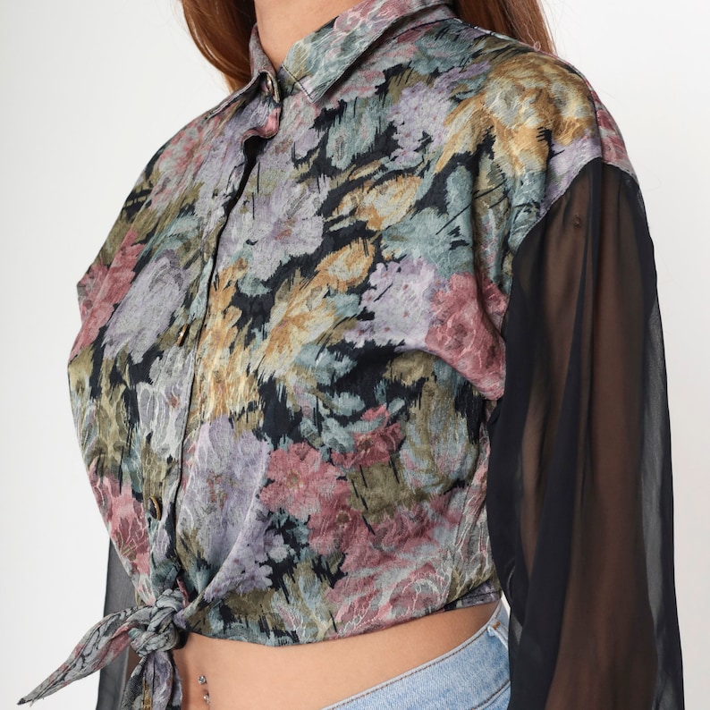 May include: A floral print button-down shirt with black sheer sleeves. The shirt has a tie-front design and a collar. The floral pattern includes shades of pink, purple, green, and gold on a black background. The sleeves are long and made of a sheer black fabric.