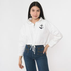 Könnte beinhalten: Weißer, kurzer Pullover mit einem marineblauen Anker-Motiv und einem Kordelzug in der Taille.