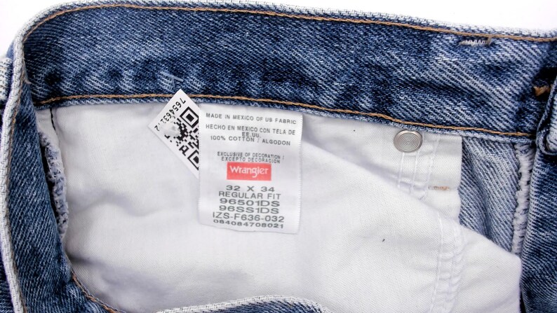 Peut inclure: Gros plan sur un jean en denim bleu avec un int&eacute;rieur blanc. Une &eacute;tiquette indique "Wrangler" avec la taille 32 x 34. Le jean est en 100% coton. L'&eacute;tiquette comprend &eacute;galement un code QR et d'autres d&eacute;tails.