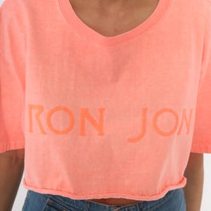 K&ouml;nnte beinhalten: Ein korallenfarbenes, kurz&auml;rmeliges, bauchfreies T-Shirt mit geripptem Rundhalsausschnitt. Auf der Vorderseite befindet sich der Schriftzug RON JON in einem etwas dunkleren Korallenton, der mittig auf der Brust platziert ist. Der Stoff ist verwaschen.