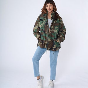 May include: A camouflage military jacket with a green, brown, and black pattern. The jacket has a zipper closure and pockets. The person is wearing light blue jeans and white sneakers. The jacket has the text "U.S. ARMY" on the right side.