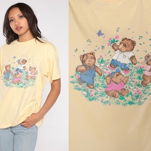 May include: A light yellow t-shirt featuring a graphic of four teddy bears in a field of flowers. The bears are wearing various outfits, including overalls and a pink shirt. Butterflies and flowers are scattered around the bears.