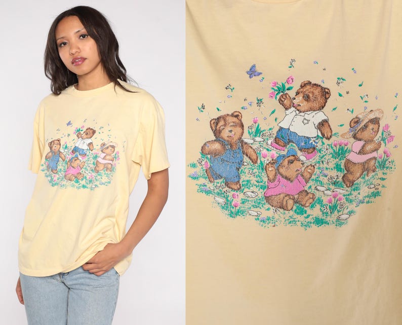 May include: A light yellow t-shirt featuring a graphic of four teddy bears in a field of flowers. The bears are wearing various outfits, including overalls and a pink shirt. Butterflies and flowers are scattered around the bears.