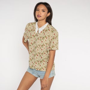 K&ouml;nnte beinhalten: Kurz&auml;rmeliges Button-Down-Shirt mit floralem Muster in Gr&uuml;n-, Rot- und Beiget&ouml;nen. Das Hemd hat einen wei&szlig;en Spitzenkragen. Das Model tr&auml;gt Jeansshorts.