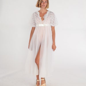 Pode incluir: Um camisão branco de gaze com detalhes em renda e um laço de fita de cetim na cintura. O camisão tem um decote em V e mangas curtas.