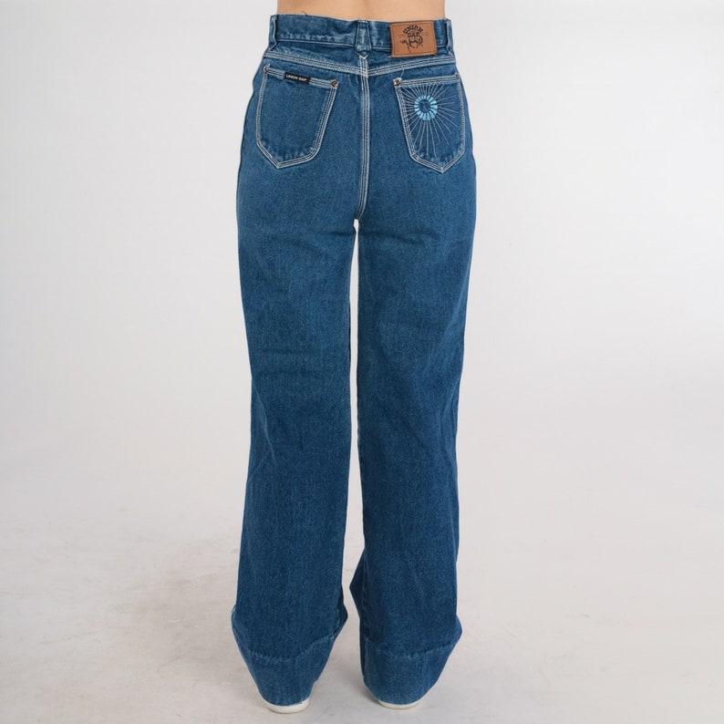 May include: A pair of high-waisted, wide-leg jeans in a dark blue wash. The jeans have a flower embroidered on the back pocket.