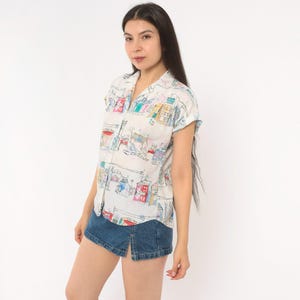 May include: A short-sleeved button-down shirt in white featuring a colorful hand-drawn style print of various buildings and houses in shades of red, blue, yellow, and green, paired with a dark blue denim mini skirt with a front slit.