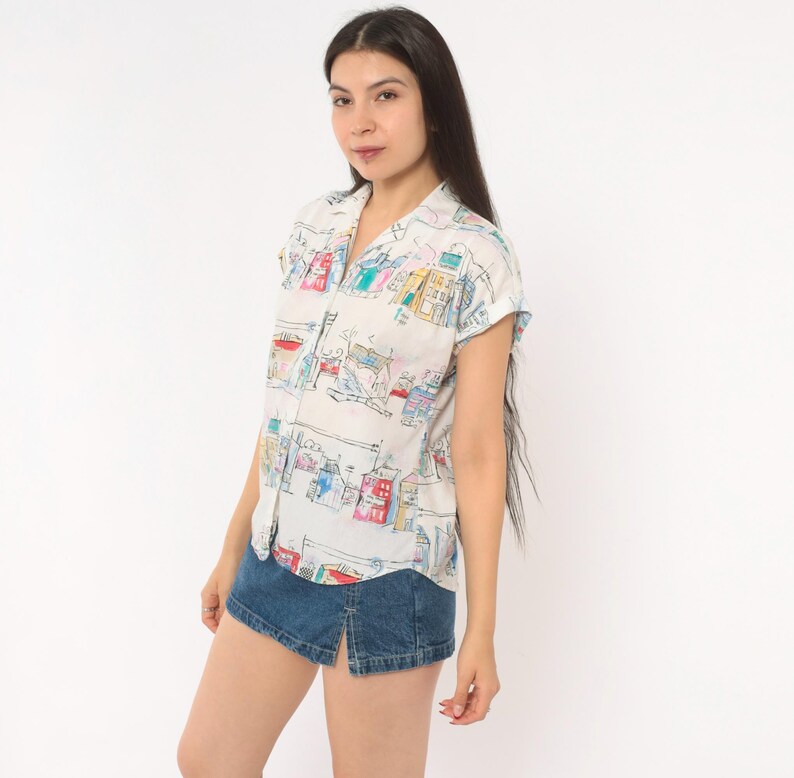 May include: A short-sleeved button-down shirt in white featuring a colorful hand-drawn style print of various buildings and houses in shades of red, blue, yellow, and green, paired with a dark blue denim mini skirt with a front slit.