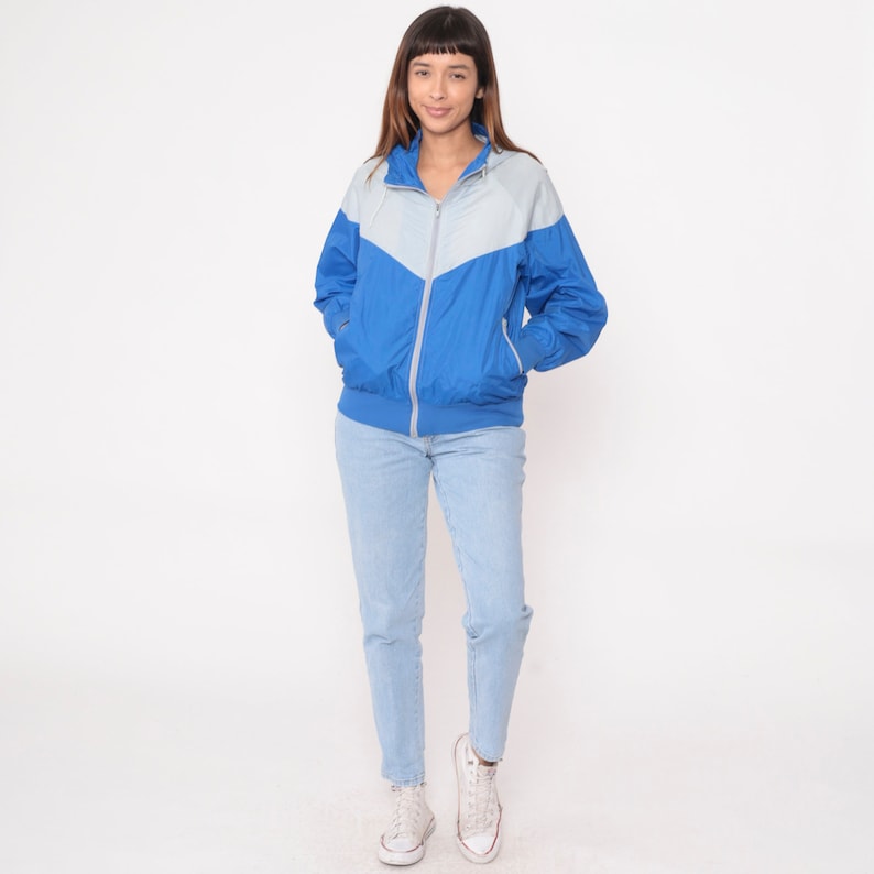 May include: A blue and gray hooded jacket with a zipper closure. The jacket has a color block design with a gray panel on the top and a blue panel on the bottom. The model is wearing light blue jeans and white sneakers.