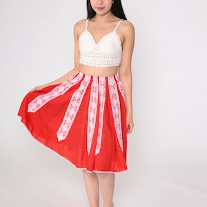 May include: A red midi skirt with white lace trim. The skirt has a flared silhouette and is made of a lightweight fabric. The model is wearing a white crochet top.