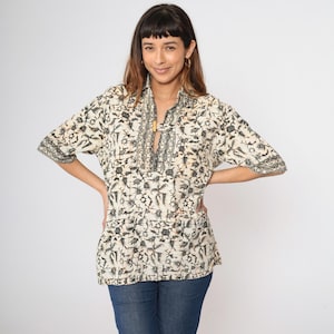 May include: Cream-colored tunic with a black floral pattern. The short-sleeved top features a mandarin collar and a decorative neckline. The tunic is paired with blue jeans. The model is smiling and has their hands on their hips.