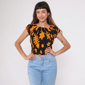 Könnte beinhalten: Schwarzes Crop-Top mit orangefarbenem Blumenmuster. Das Oberteil hat einen quadratischen Ausschnitt und kurze Ärmel. Das Model trägt eine hellblaue Jeans. Das Oberteil ist aus einem strukturierten Stoff gefertigt.