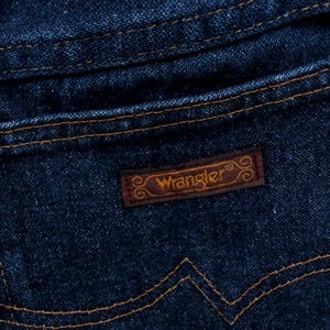 May include: Close-up of a dark blue denim jeans pocket. The pocket features a brown leather patch with the brand name "Wrangler" in a stylized font. The stitching is a contrasting tan color.
