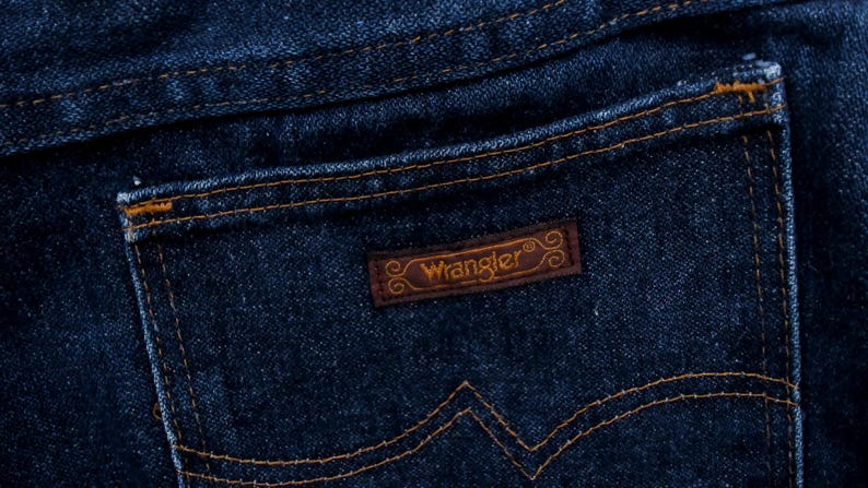 May include: Close-up of a dark blue denim jeans pocket. The pocket features a brown leather patch with the brand name "Wrangler" in a stylized font. The stitching is a contrasting tan color.
