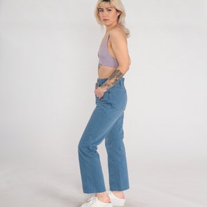 Op de afbeelding: Een vrouw draagt een lichtpaarse crop top en een blauwe denim jeans. Ze staat met haar hand in haar zak en kijkt naar de camera.