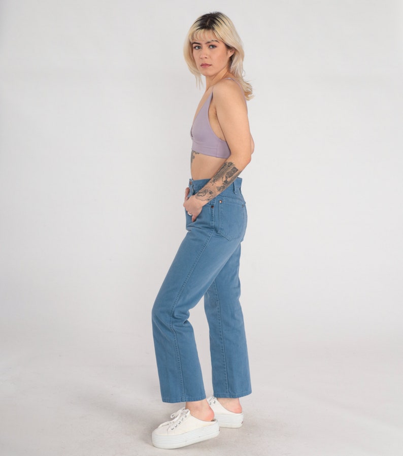 Op de afbeelding: Een vrouw draagt een lichtpaarse crop top en een blauwe denim jeans. Ze staat met haar hand in haar zak en kijkt naar de camera.