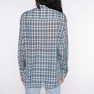Op de afbeelding: Een geruit shirt met lange mouwen in groen, marineblauw en wit. Het shirt heeft een klassiek ontwerp met knopen en een traditioneel ruitpatroon. De achterkant van het shirt is zichtbaar en toont de textuur en kleurvariaties van de stof.