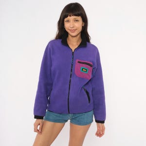 May include: A purple fleece zip-up jacket featuring a black collar, black cuffs, and a contrasting magenta chest pocket with a black zipper. The pocket displays a rectangular patch with the text REI in orange and teal lettering on a black background.