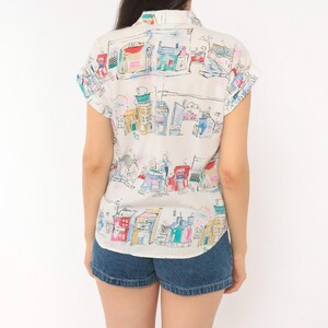 May include: A short-sleeved button-down shirt in an off-white color featuring a colorful hand-drawn style print of various buildings and houses in shades of blue, red, yellow, and green, paired with dark blue denim shorts with a cuffed hem.