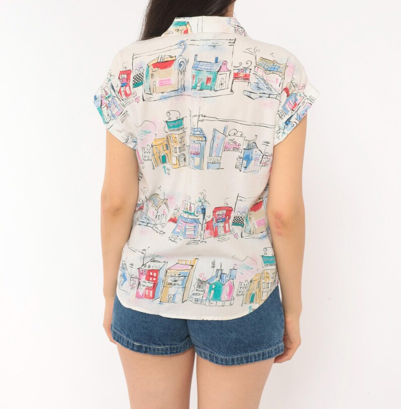 May include: A short-sleeved button-down shirt in an off-white color featuring a colorful hand-drawn style print of various buildings and houses in shades of blue, red, yellow, and green, paired with dark blue denim shorts with a cuffed hem.